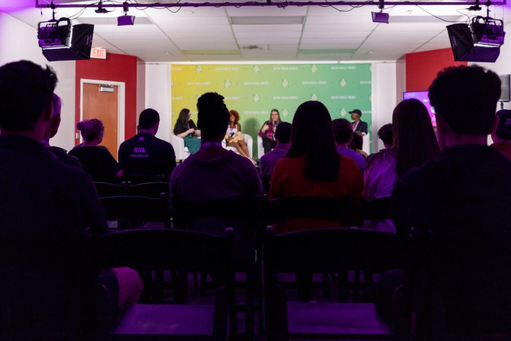 An audience sits in a purple lit breakout room listening to a panel of presenters. 