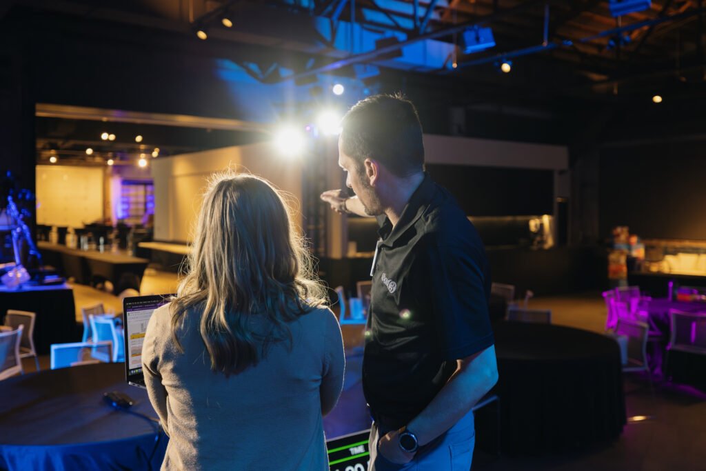 A production manager discusses a room layout during setup for an event with a client. The room is brightly lit with blue, purple, and daylight lights. 