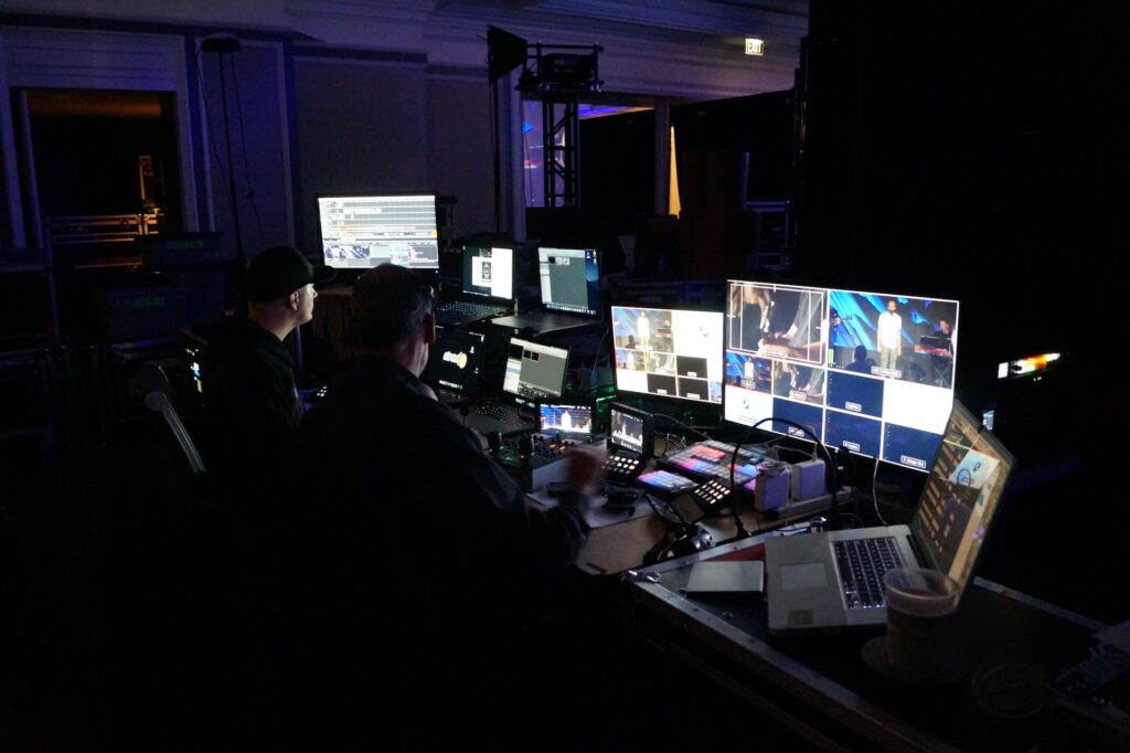 2 production techs sit in front of an array of screens in a dark room.