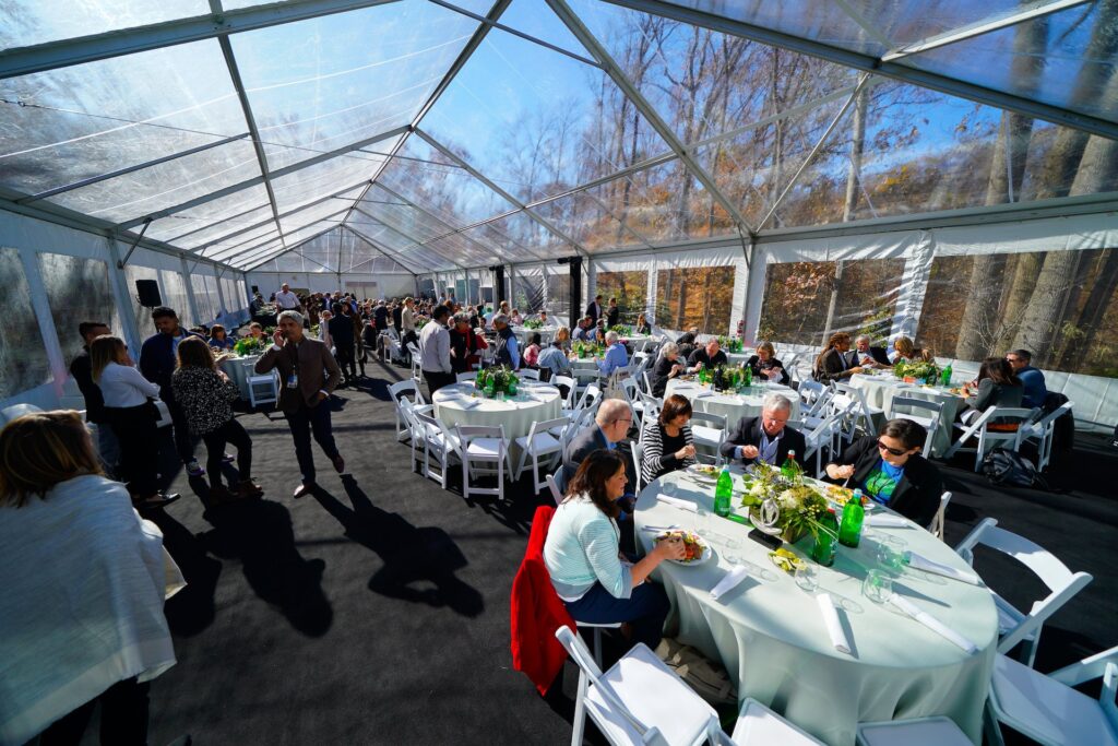 An enclosed outdoor tent surrounds hundreds of tables with guests eating. 
