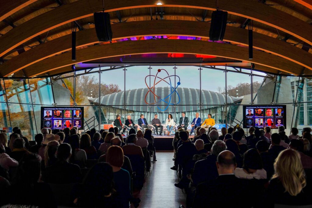 Hundreds of event attendees sit behind a stage with multiple presenters. Light fills the ceiling of a glass wall venue.