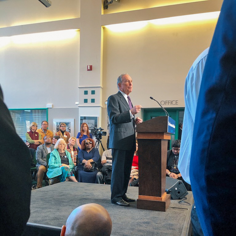 Mike Bloomberg stands and speaks at a podium to hundreds of guests at an indoor event.