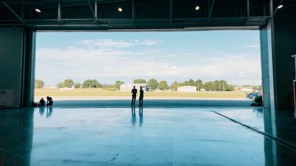 Two men stand at the middle of a massive airplane hangar door in daytime.
