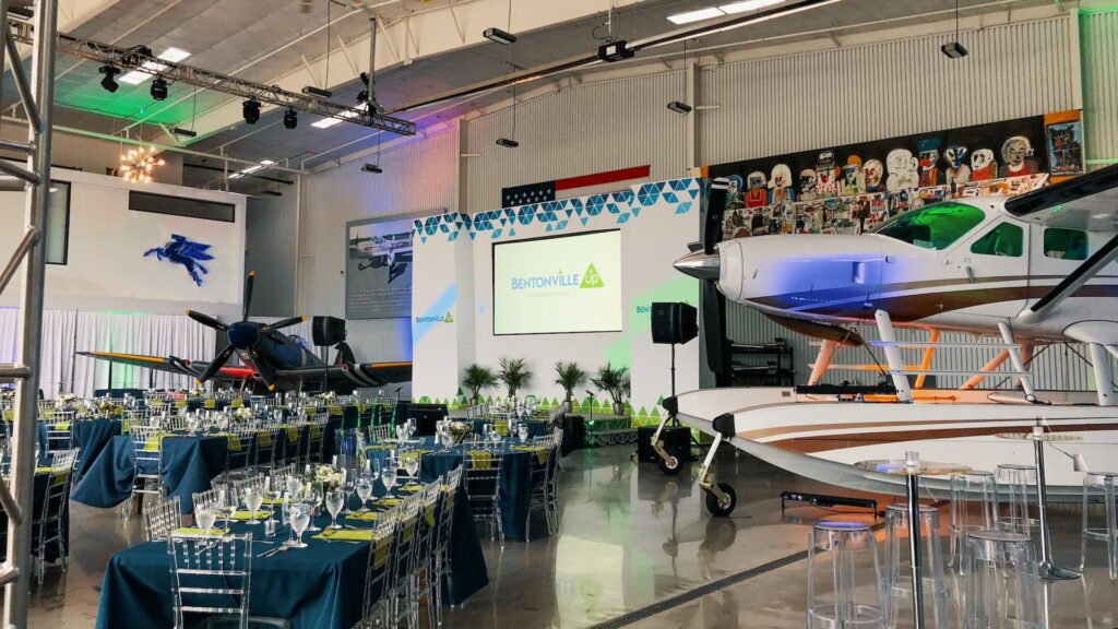 A screen is accompanied by two airplanes and many tables in an airplane hangar.