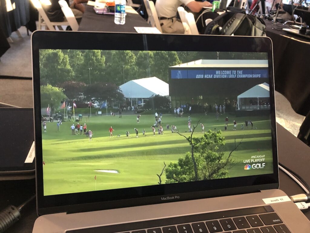 A computer monitor shows a livestream with bright green grass and golf athletes performing