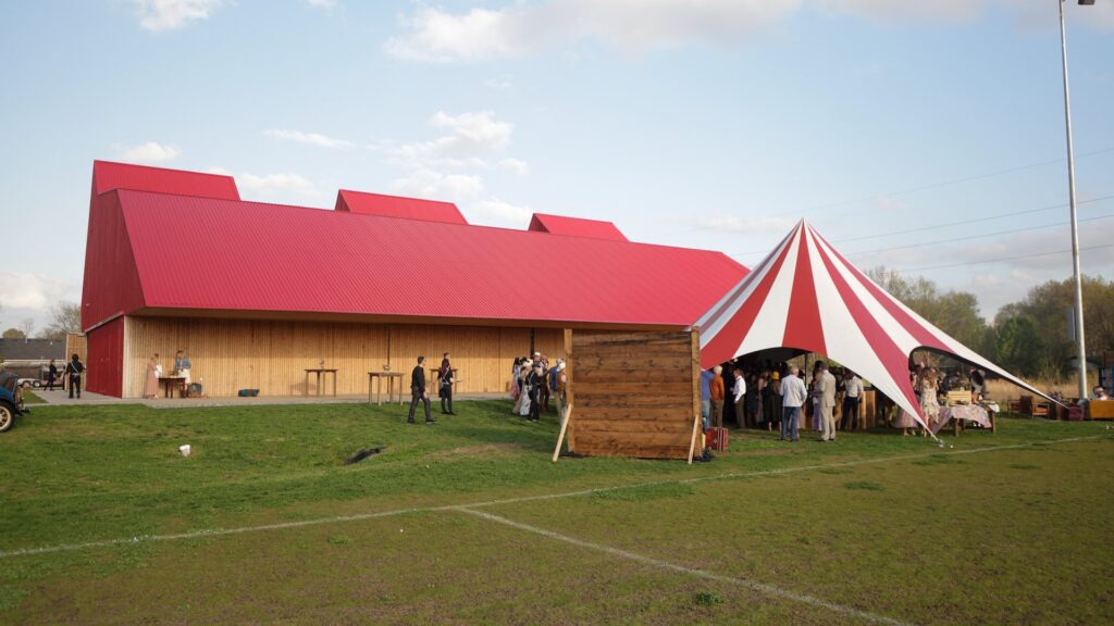 Red barn in a field with a large crowd standing outside.