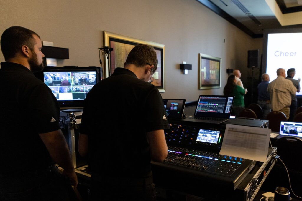 An audio technician wearing all black sits in the back of a crowded room mixing audio for an event. 
