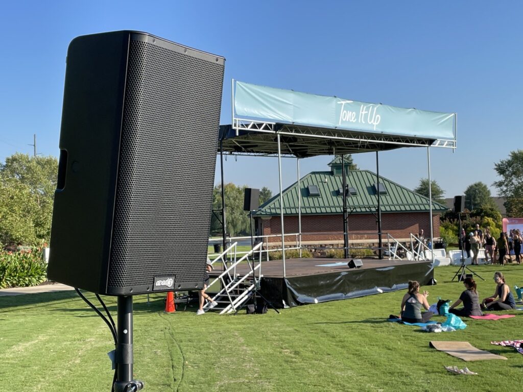 A loudspeaker sits next to a portable outdoor stage on a summer day.