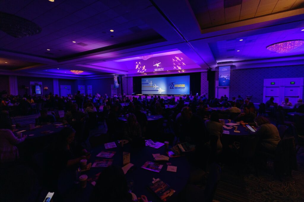 A purple lit room with hundreds of attendees. 
